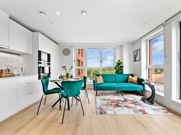 Bright living room with white kitchen on the left, teal sofa with pillows, four teal dining chairs around a black table, large windows with city and greenery views, colorful area rug, and minimal decor.