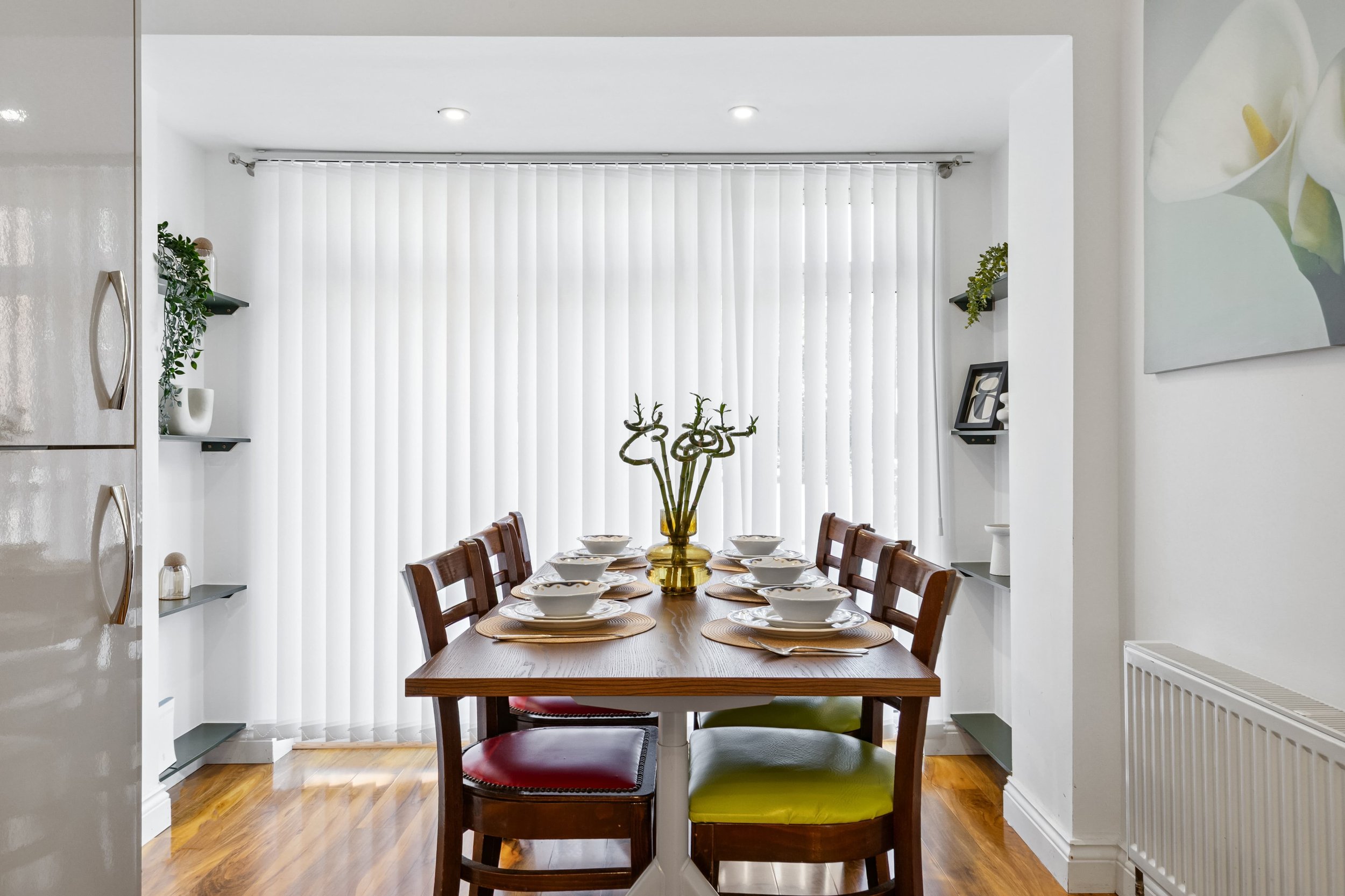 Dining room with a wooden table set with plates, bowls, and a yellow vase with decorative branches, surrounded by mixed colored chairs in front of white vertical blinds.