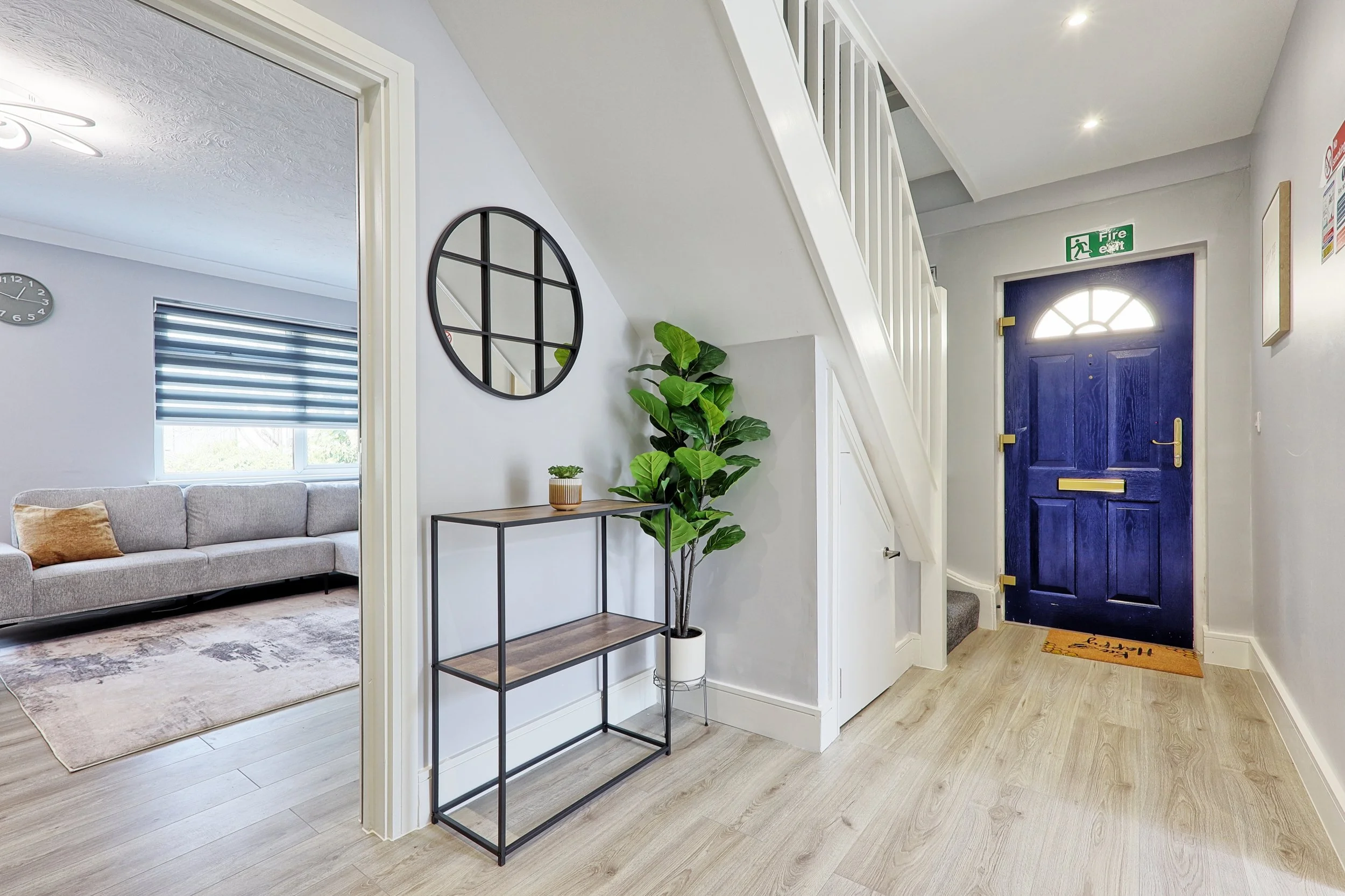 Entry hallway with a bright blue front door, a staircase, a potted plant, and a console table with a small plant. To the left is a doorway leading to a living room with a sofa and window blinds.