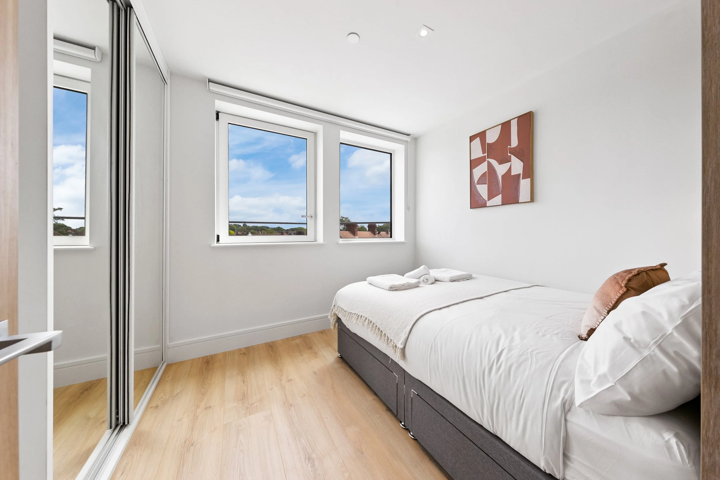 A minimalistic bedroom with a double bed, white linens, and a brown pillow. There is a large window showing a blue sky with clouds, and a piece of abstract wall art. A sliding closet with mirrored doors is on the left, and the room has light wooden flooring.