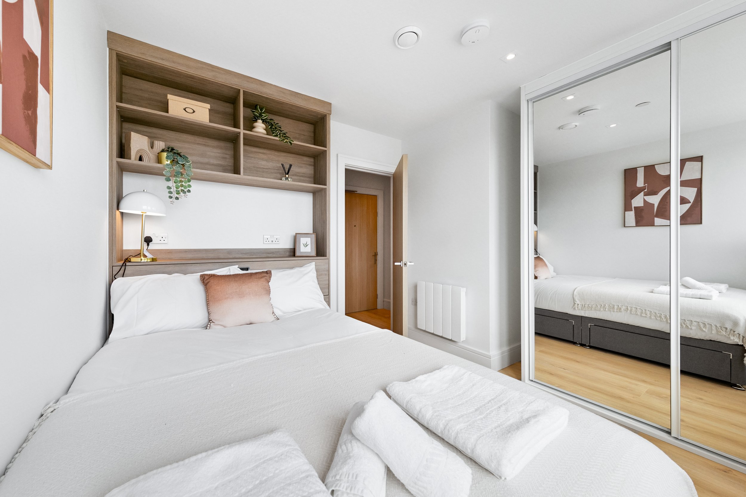 A modern bedroom with a neatly made bed, wooden shelving above the headboard, a mirrored closet door, artwork on the walls, and neatly folded towels on the bed.