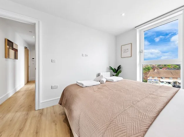 A bedroom with a bed covered in a beige quilt, white pillows, and folded towels on top. There is a large window showing a view of houses and a blue sky with clouds. The room has white walls, light wood flooring, and a framed picture on the wall beside the window.