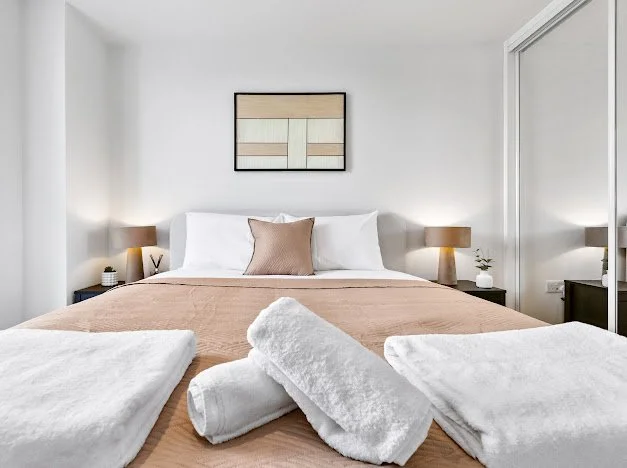 Bedroom with neatly made bed, white bedding, a beige throw blanket, and two towels in front. Two nightstands with lamps and small plants, and a framed artwork on the wall.