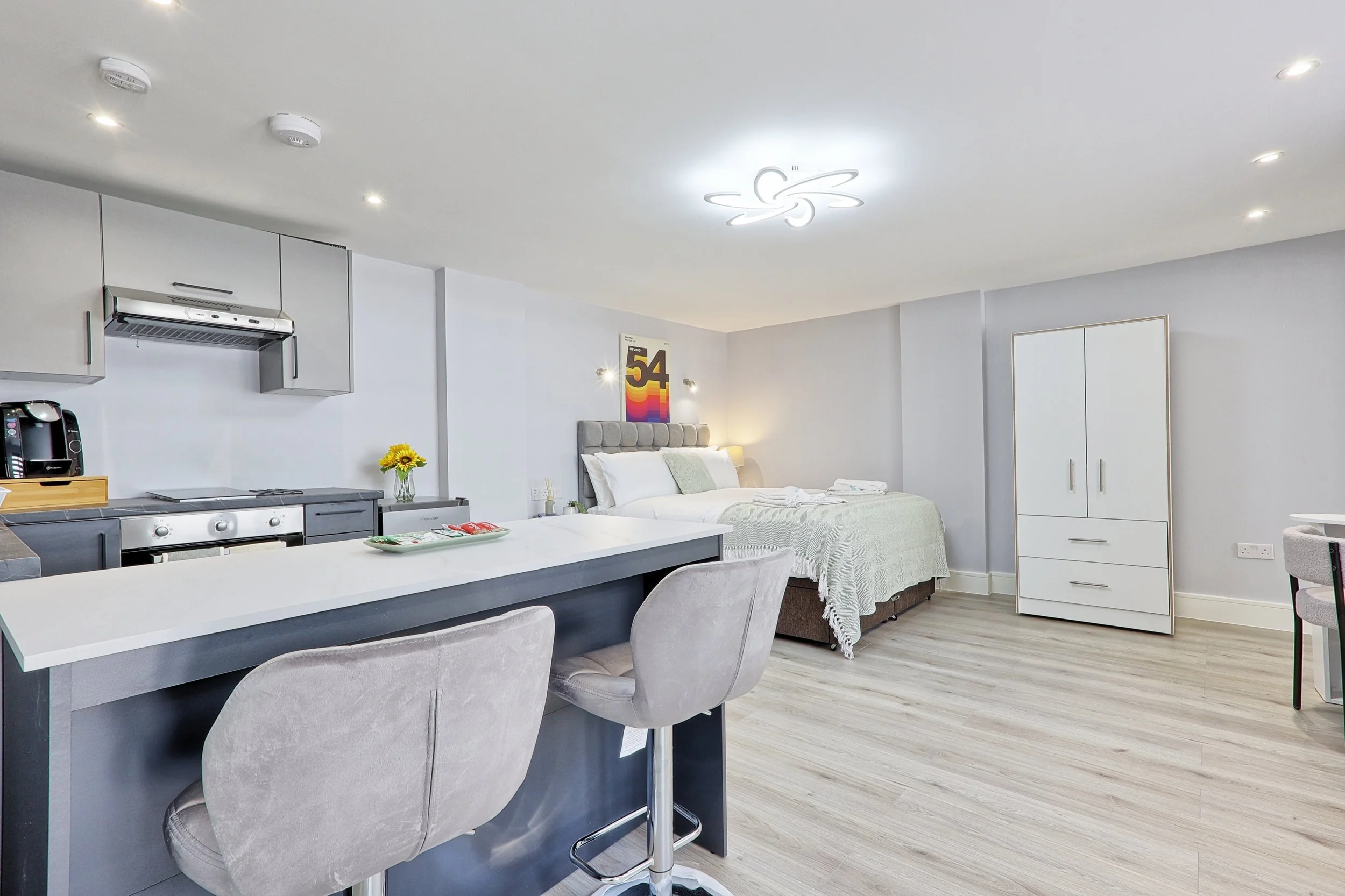 Modern studio apartment with a combined kitchen and sleeping area. The kitchen has a stove, oven, microwave, and a small countertop with a sunflower in a vase. The sleeping area features a bed with white and gray bedding, a headboard, and a vibrant "54" poster on the wall. The space has light-colored wooden floors and white walls, with built-in ceiling lighting and a minimalist decor.