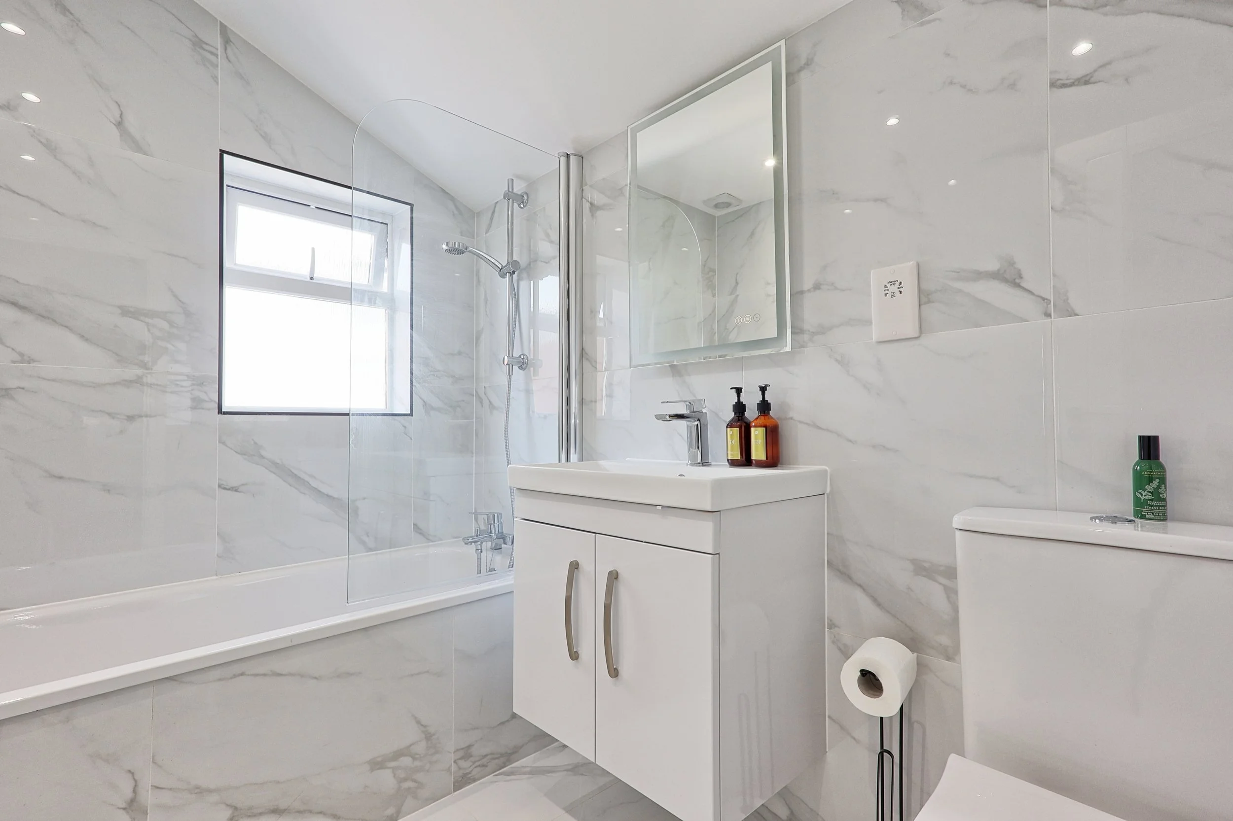 Modern bathroom with marble walls and floor, white bathtub with glass shower partition, small white vanity with two bottles on top, large mirror, toilet with toilet paper roll, window on wall, and toiletries on toilet tank.