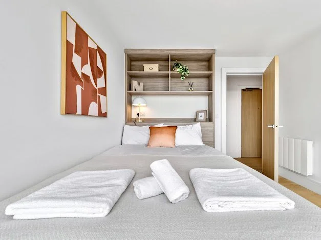 A neatly made bed with white bedding and an orange pillow, with rolled towels on top, in a bedroom featuring a wooden headboard, open shelving with decor, a framed picture, and a bedside lamp, with an abstract painting on the wall and a door leading to another room.