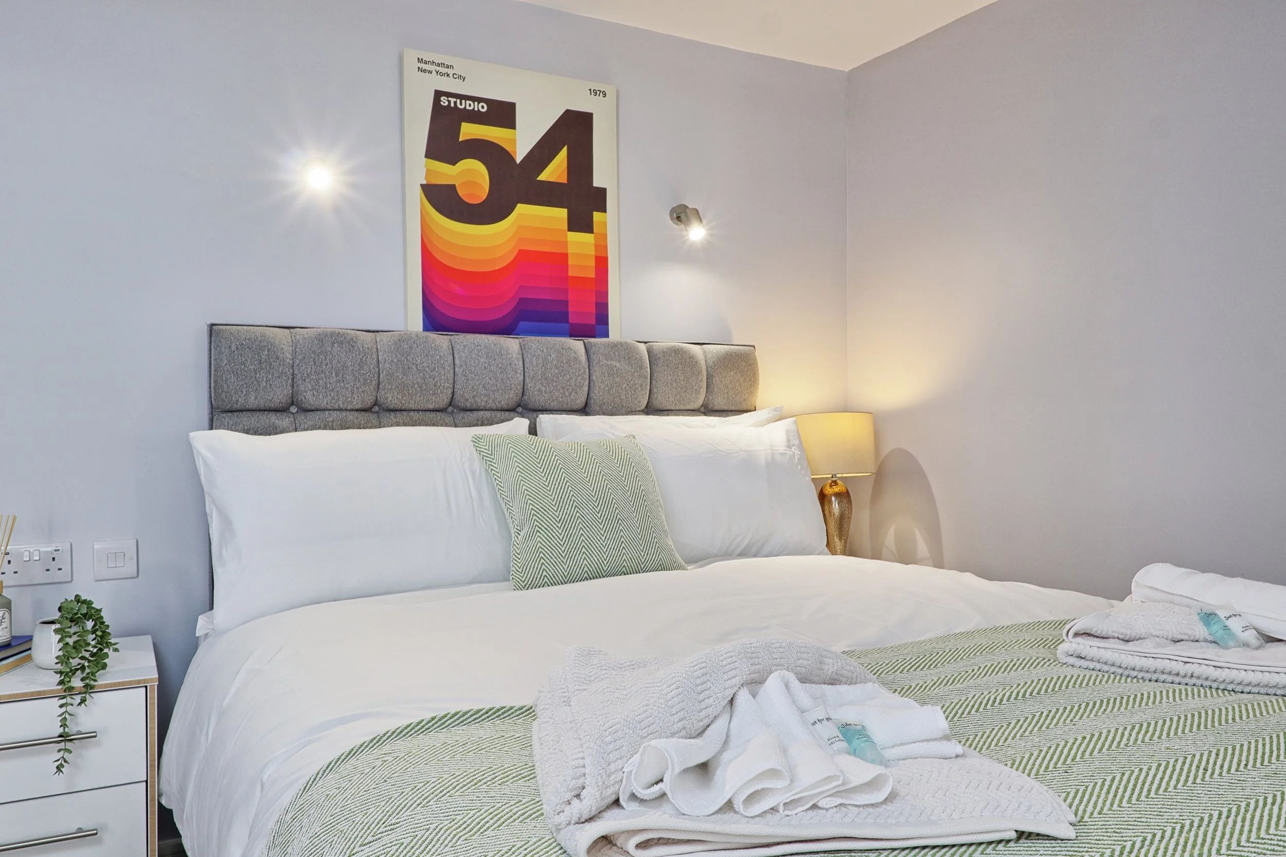 A neatly made bed with white bedding, colorful striped pillow, and a green patterned throw blanket in a bright bedroom. A wall-mounted reading light, a decorative table lamp, and a framed colorful abstract poster are visible on the wall.