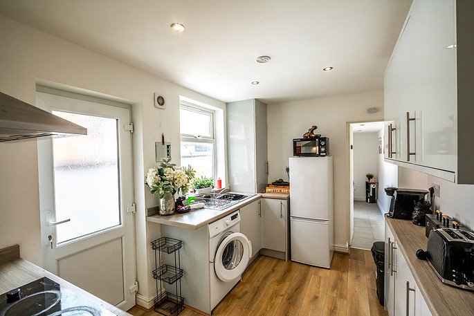 Kitchen with white cabinets, a refrigerator, microwave, washing machine, and various appliances, with hardwood floors and natural light from windows.