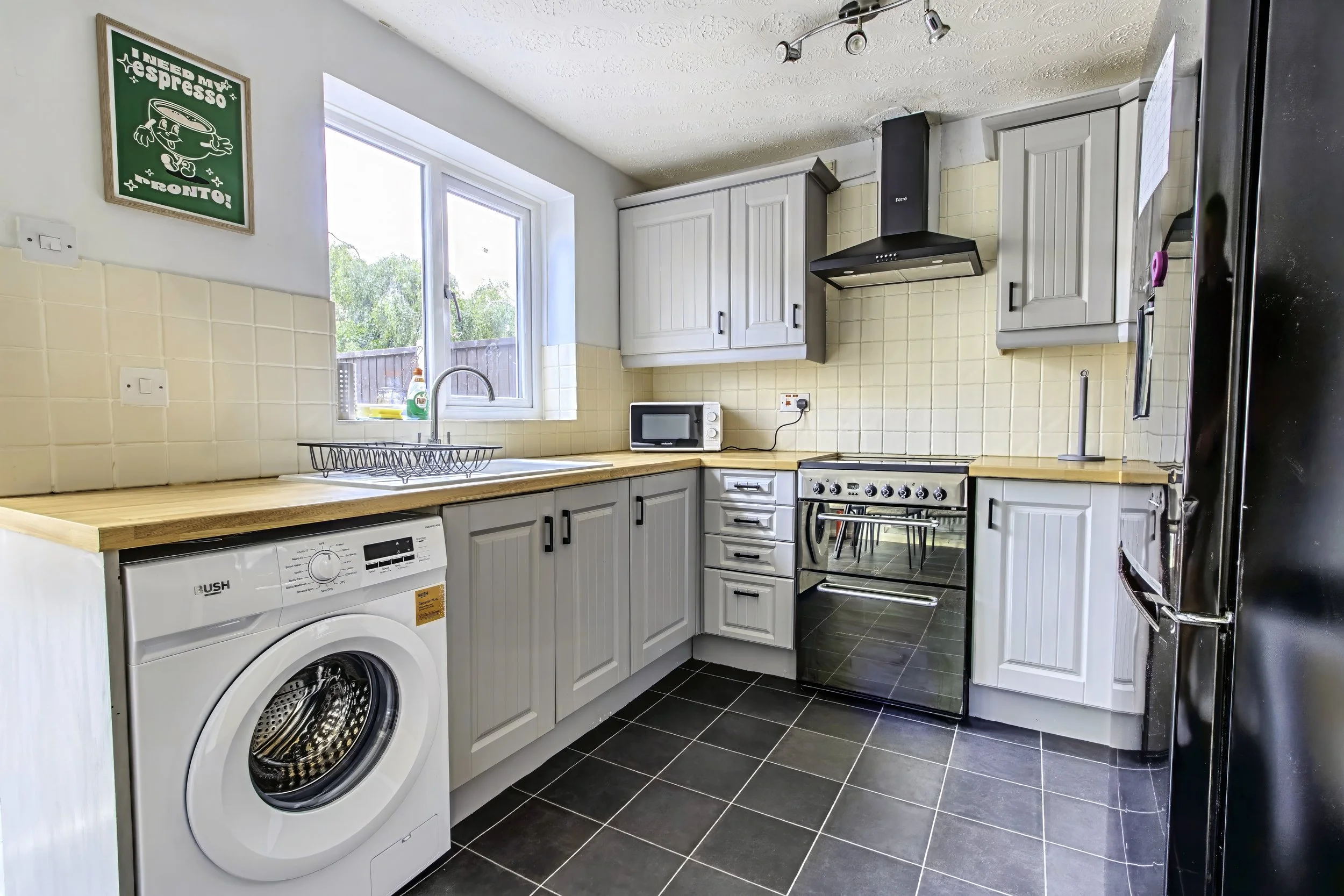 Kitchen with white cabinets, a refrigerator, microwave, washing machine, and various appliances, with hardwood floors and natural light from windows.