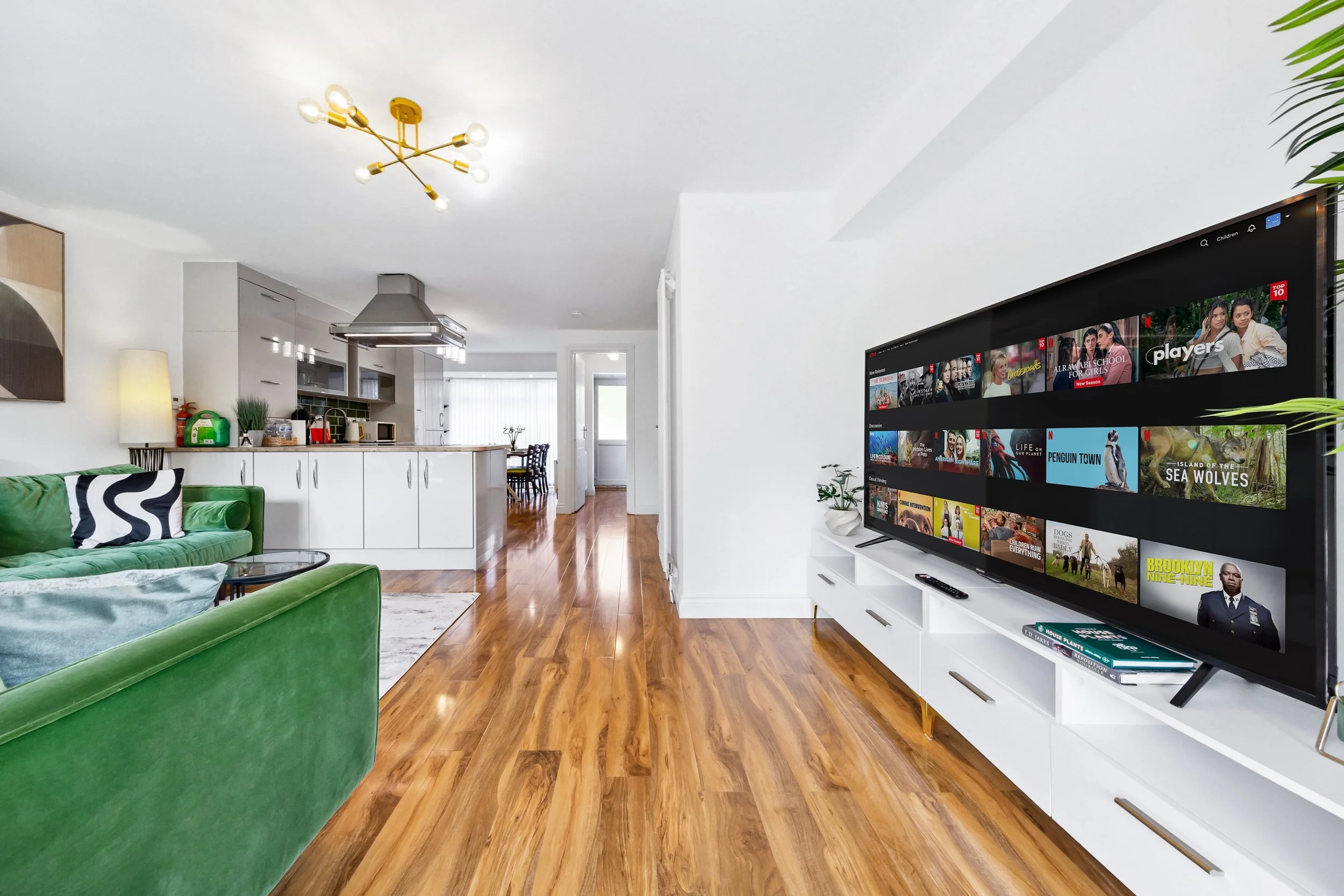 Living room with hardwood floors, white walls, a green velvet sofa, a large flat-screen TV displaying a streaming service, a white TV stand, and a kitchen area in the background with a dining table.