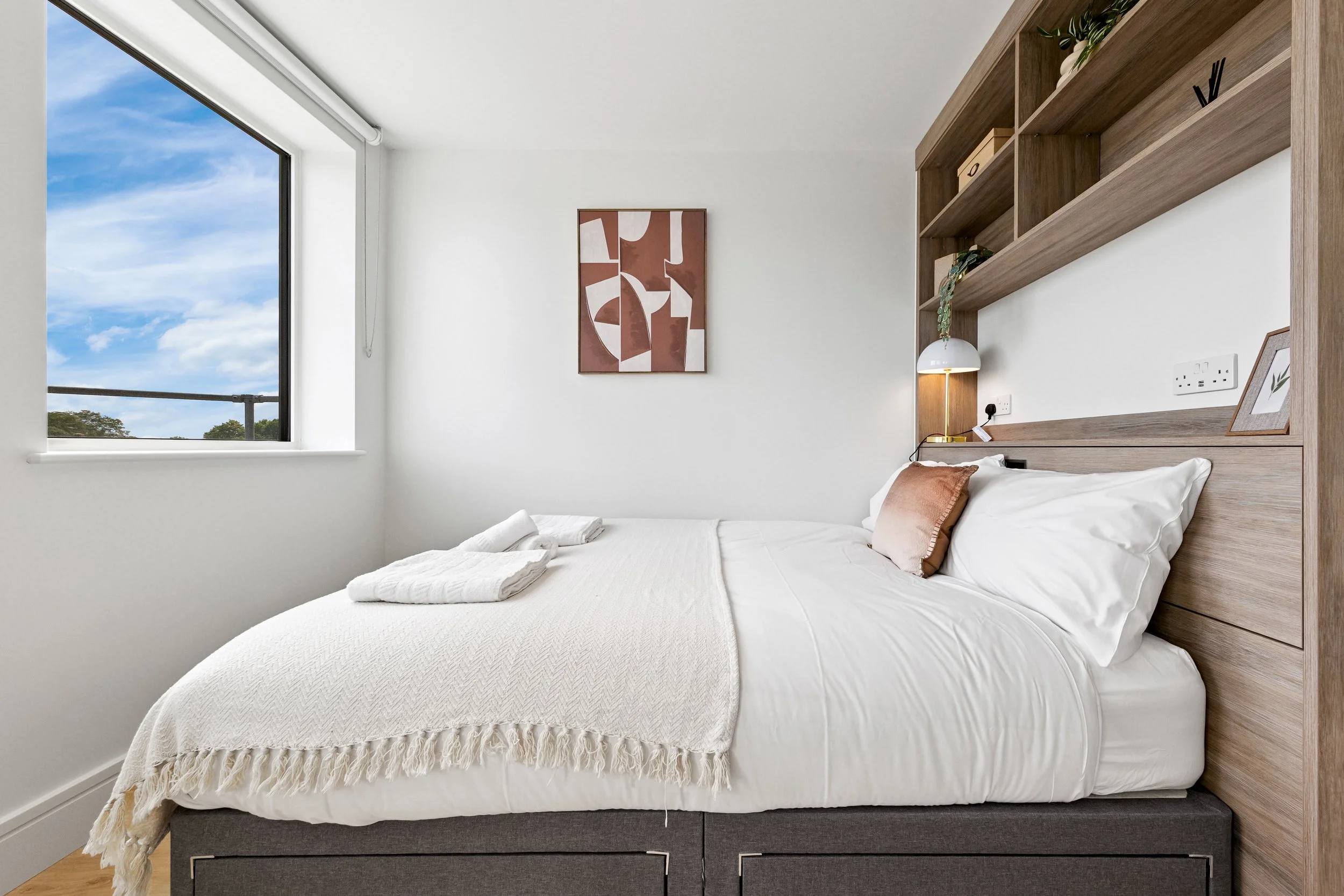 Modern bedroom with white walls, a large window showing a blue sky with clouds, a bed with white linens, a pink decorative pillow, and a beige throw blanket. There is a wooden headboard with built-in shelves, a small desk lamp, and framed art on the wall and shelf.