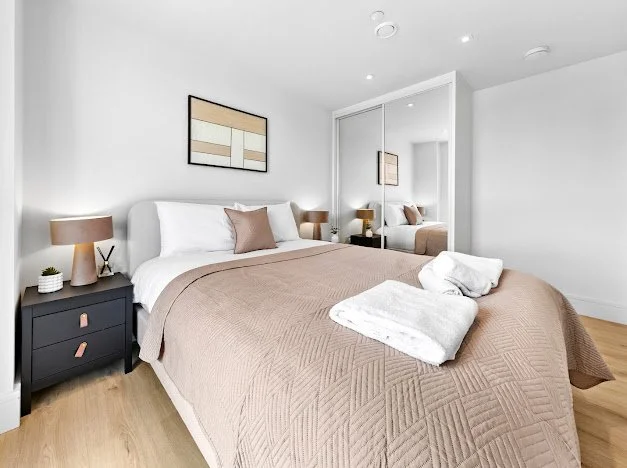 Modern bedroom with a beige bedspread, white pillows, and a black nightstand with a lamp, small plant, and decorative item. Mirrored closet doors and minimal wall art are visible.