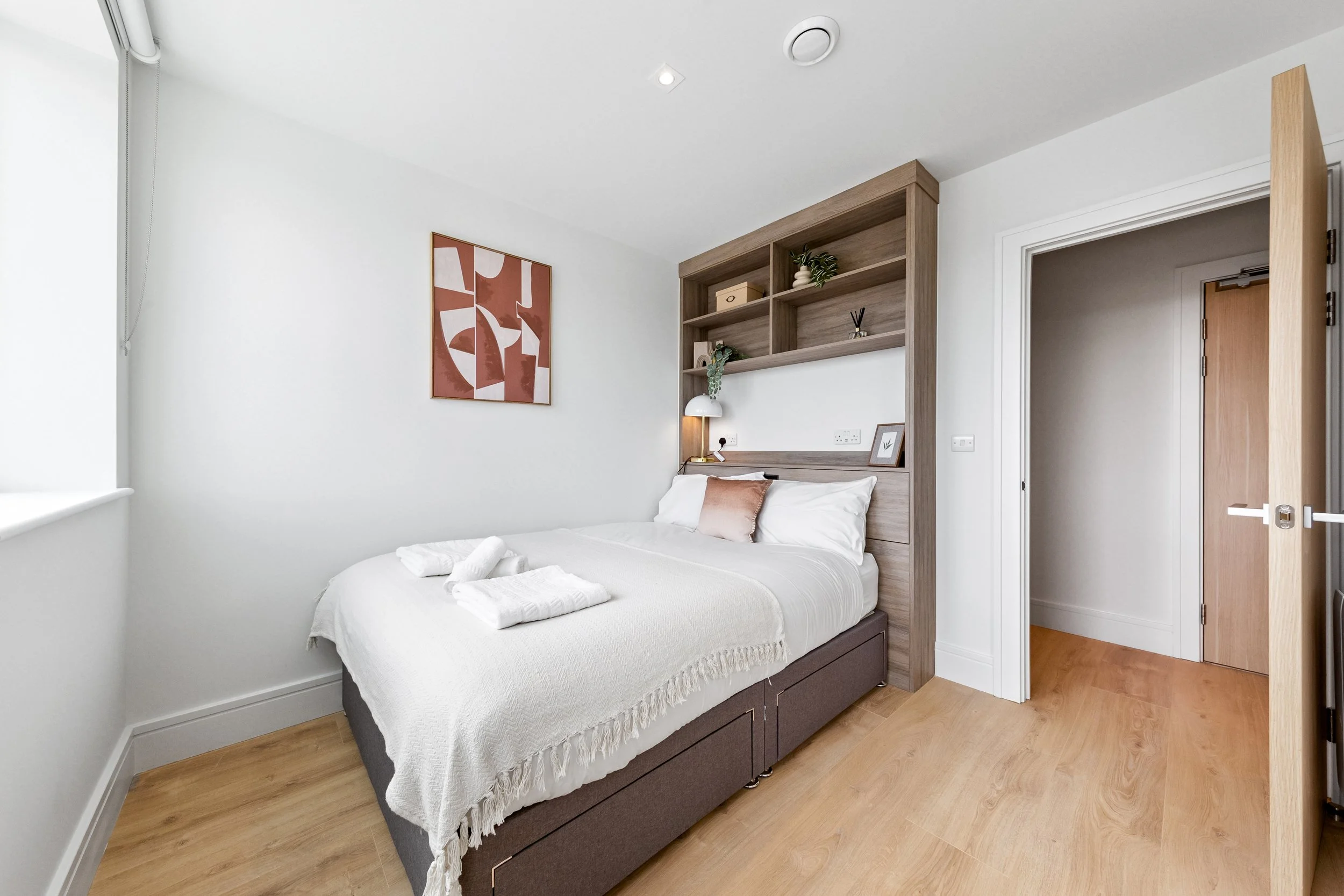 A modern bedroom with a bed, white linens, and a brown decorative pillow. Wooden flooring and a built-in bookshelf serve as a headboard, with a small lamp, framed picture, and decorative items. The room has white walls, a window with a blind, and an open door leading to another room.