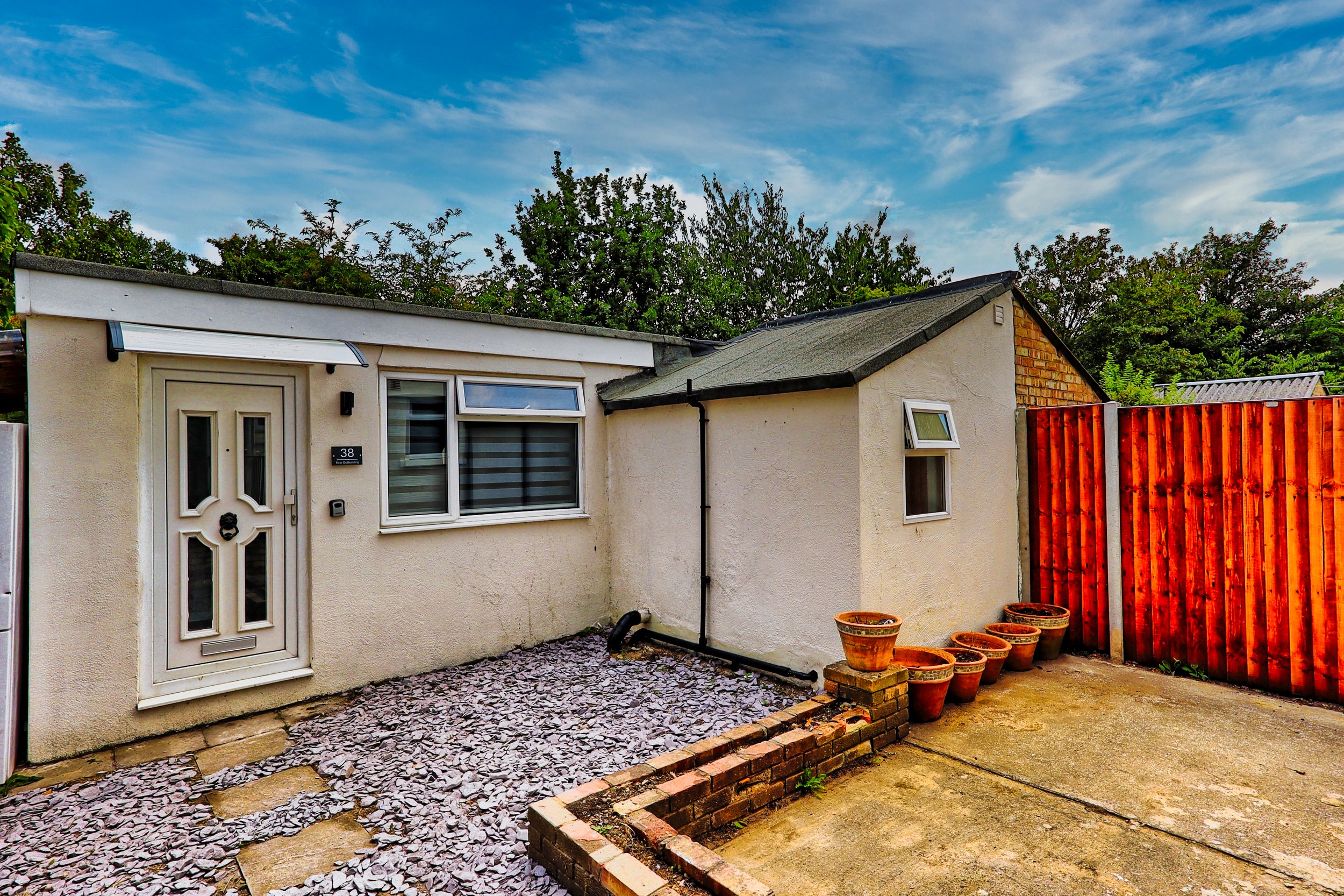 Backyard view of a small house with a white exterior, two small windows, a white front door, a gravel area, a concrete patio, a red wooden fence, and five flower pots around the corner of the house.