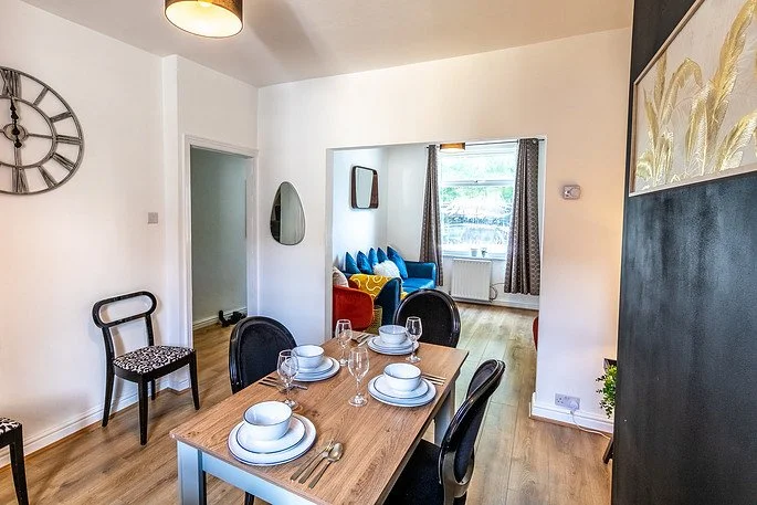Dining room with a wooden table set for four, black chairs, and a view into a living room with a blue sofa, cushions, and a window.