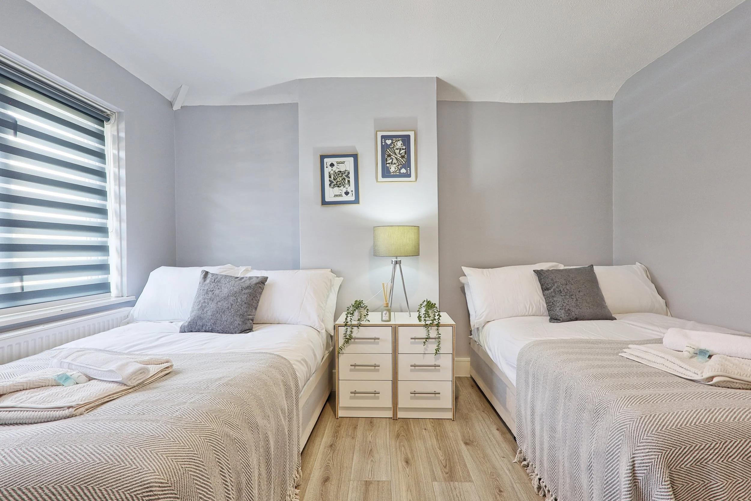 Two neatly made beds with white bedding and gray pillows, separated by a small white nightstand with a lamp and decorative plants, in a bedroom with gray walls, a window with striped blinds, and framed artwork on the wall.