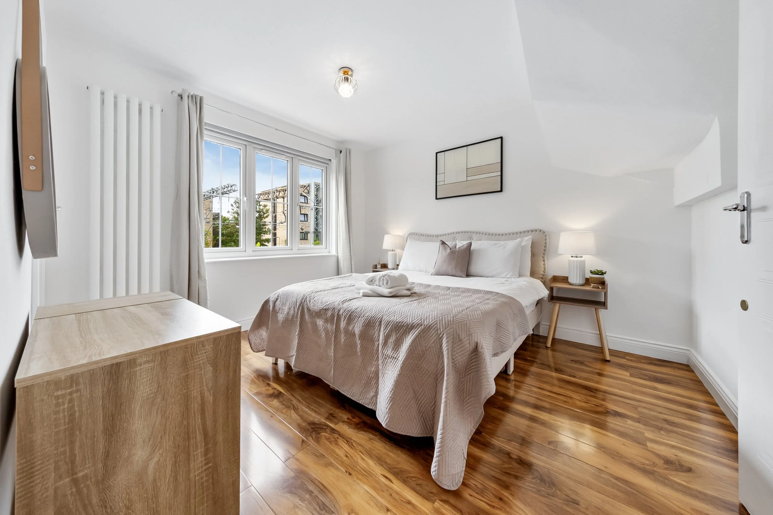 Modern bedroom with white walls, wooden flooring, a bed with beige and white bedding, two bedside tables with lamps, a window with curtains, and a piece of abstract art above the bed.