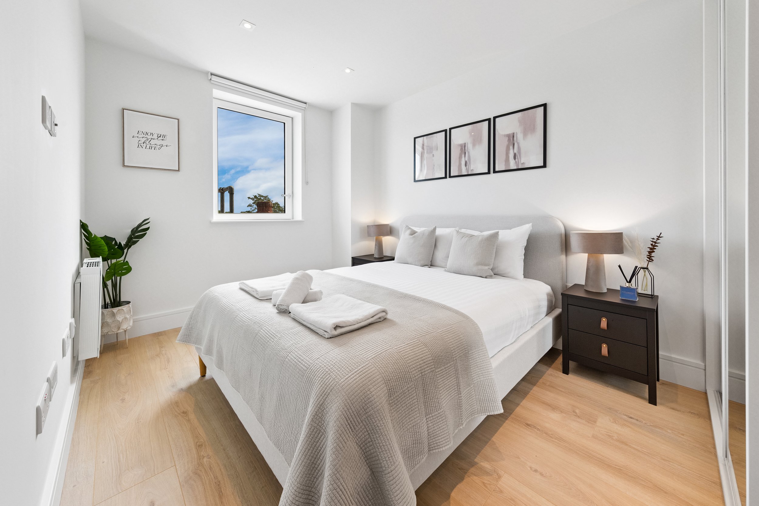 Minimalist bedroom with a white bed, beige quilt, and folded towels on top. Two nightstands with lamps and decorative items. Artwork on the wall above the bed. A window showing trees and sky, and a potted plant near the corner.