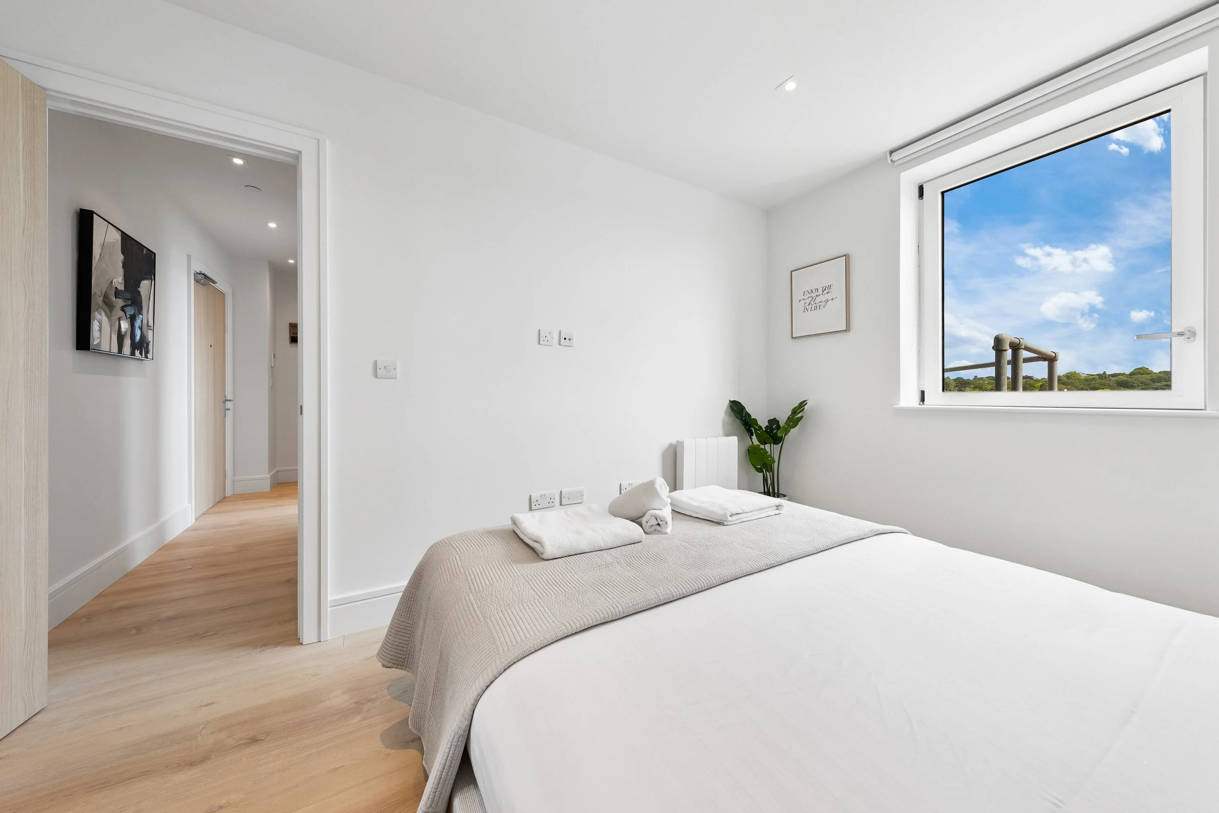 A minimalist bedroom with white walls, a large window showing blue sky and greenery, a bed with white linens, and a small plant in the corner.