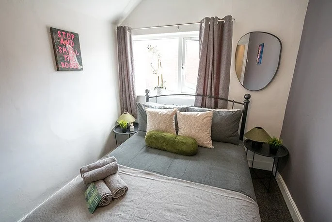 A neatly made bed with gray and white bedding, two beige pillows, a green bolster pillow, and rolled towels on the foot of the bed in a small, well-lit bedroom. The room has a window with curtains, a round mirror on the wall, two black bedside tables with lamps and plants, and a pink sign on the wall.