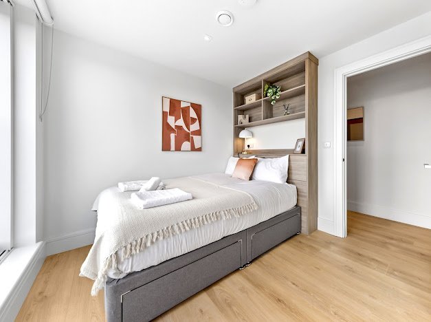 A bedroom with a bed, white bedding, a headboard with built-in shelves, wall art, and hardwood flooring.