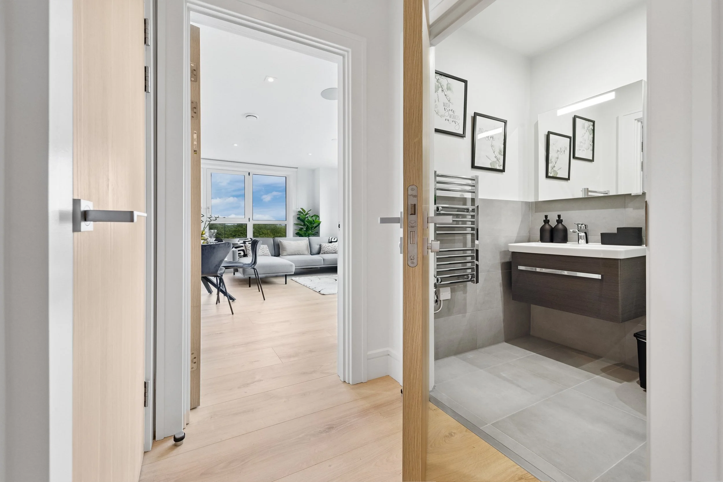 View from hallway showing a bathroom on the right with a sink, mirror, and black decor, and a living room with a large window, gray sofa, and dining table in the background.