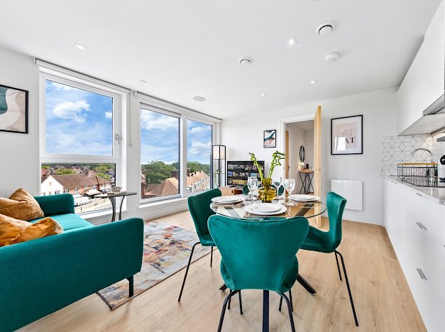 Bright, modern open-concept living and dining area with large windows showing a neighborhood view, a turquoise sofa, a round dining table with teal chairs, and a white kitchen with geometric tile backsplash.