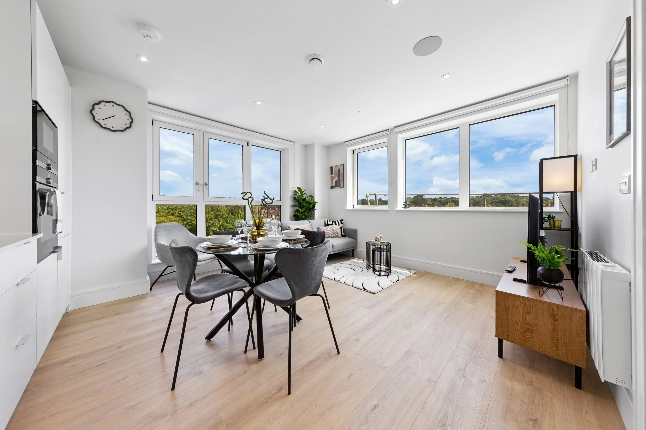 Bright living room with large windows, wooden floor, dining table with five chairs, sofa, flat-screen TV, and modern decor.