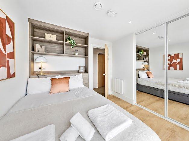 Modern bedroom with a bed, decorative pillows, a built-in shelf with accessories, and mirrored closet doors reflecting the room.
