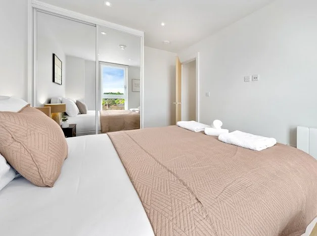 A bedroom with a bed covered in a beige quilt and pillows, white towels on top, a large mirror door closet reflecting a window and outside view, and a white wall with a door and light switches.