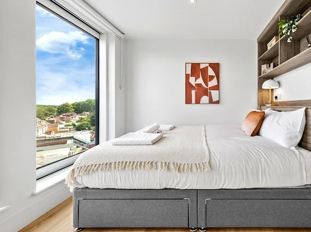Modern bedroom with a large window showing a cityscape and greenery, a neatly made bed with white linens, a wooden headboard with built-in shelves, a small lamp, and abstract wall art.