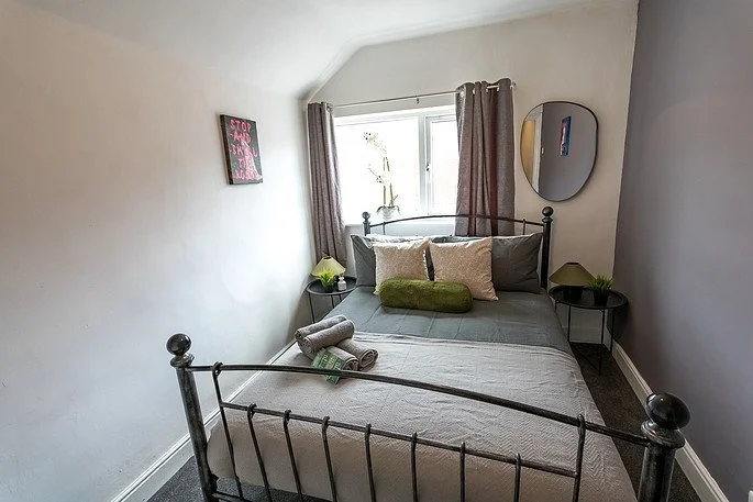 A small bedroom with a metal bed frame, gray bedding, and decorative pillows, including a green bolster. There are two small bedside tables with plants, a window with curtains, a mirror on the wall, and a small framed sign on the wall that says 'STOP THINKING, START DOING'. Two rolled towels are on the bed.