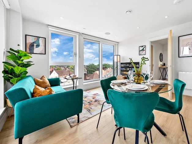 Bright living room with teal sofas, a round dining table set with white plates and glassware, large windows showing a view of houses and trees, and modern wall art.