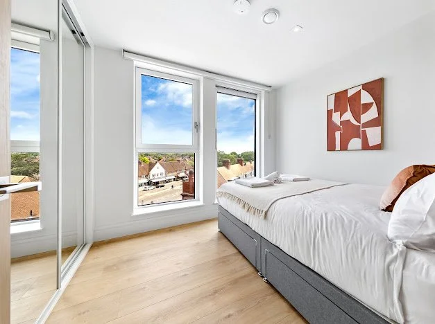 Bright bedroom with white walls, large windows showing a neighborhood view with blue sky, a bed with white linens, and a modern abstract painting on the wall.