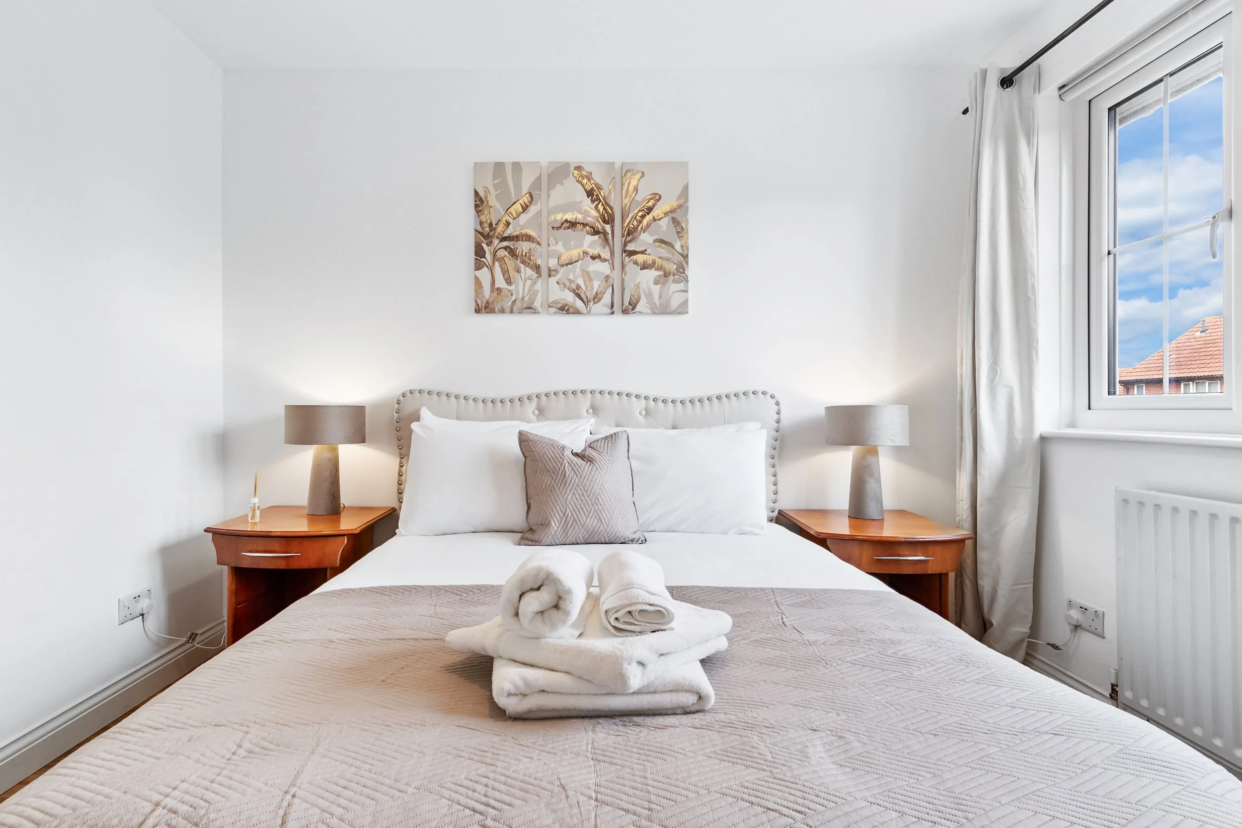 A neatly made bedroom with a white bed, beige quilt, and decorative pillow, flanked by two wooden nightstands with gray lamps. There are folded towels on the bed, a three-panel artwork of banana leaves above the headboard, a window with white curtains showing rooftops and a blue sky outside.
