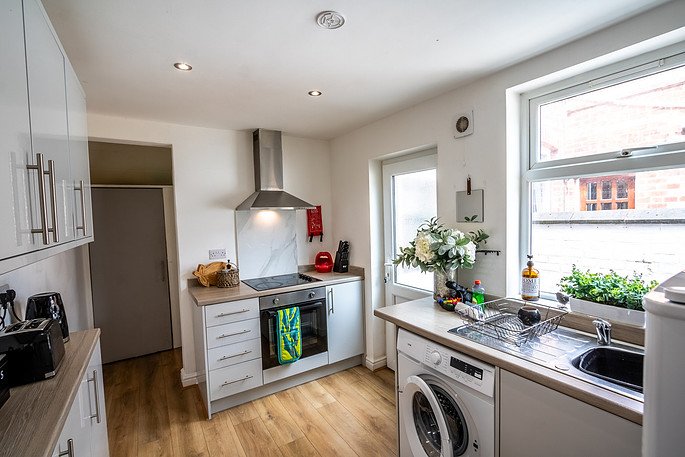 Kitchen with white cabinets, stainless steel stove and exhaust hood, window with potted plants, washing machine, and various kitchen items.