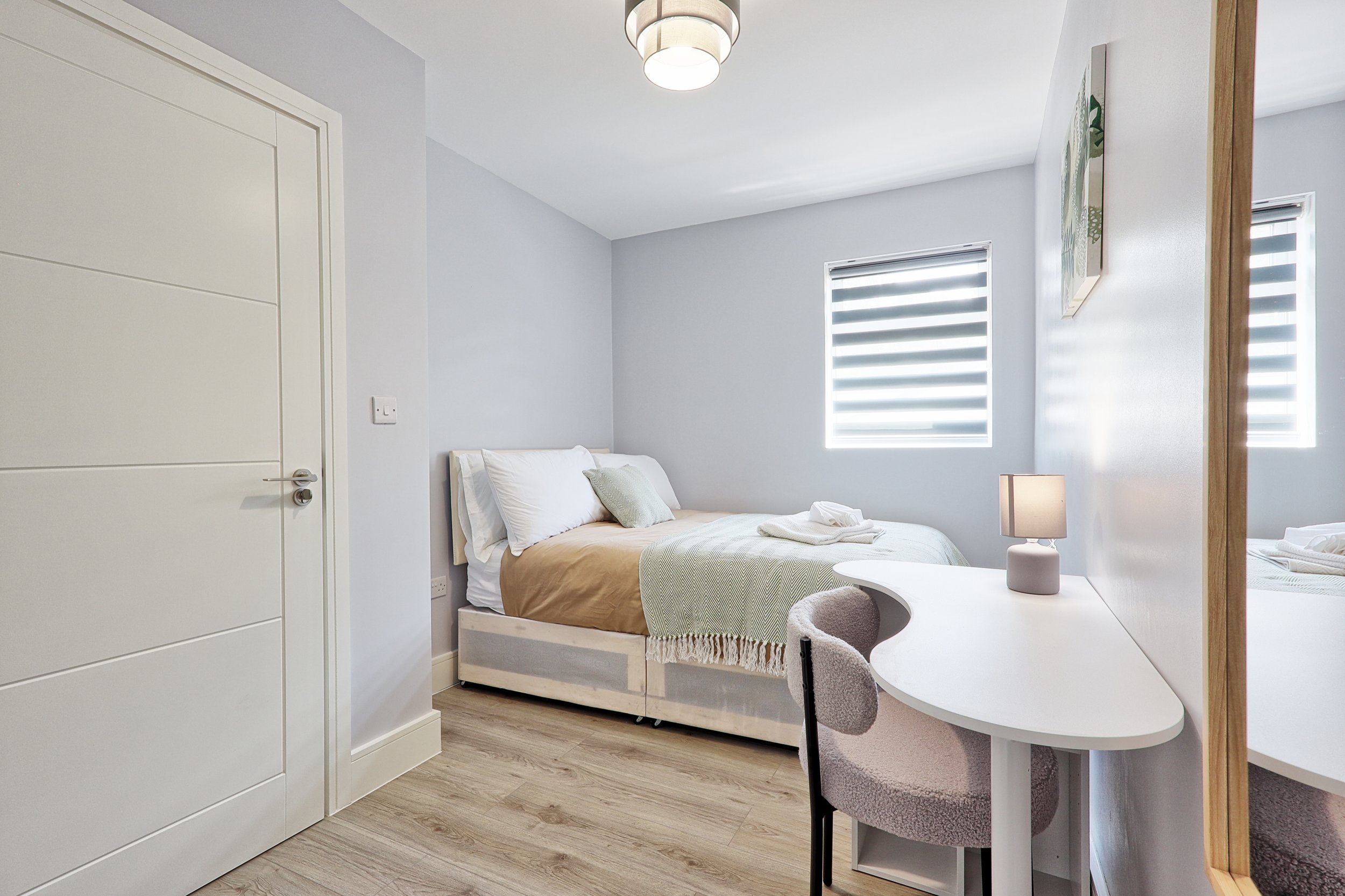 Sunlight streams through two windows with zebra blinds into a modern bedroom with light gray walls, a white door, a bed with beige bedding, a small white desk with a beige chair, and a lamp.