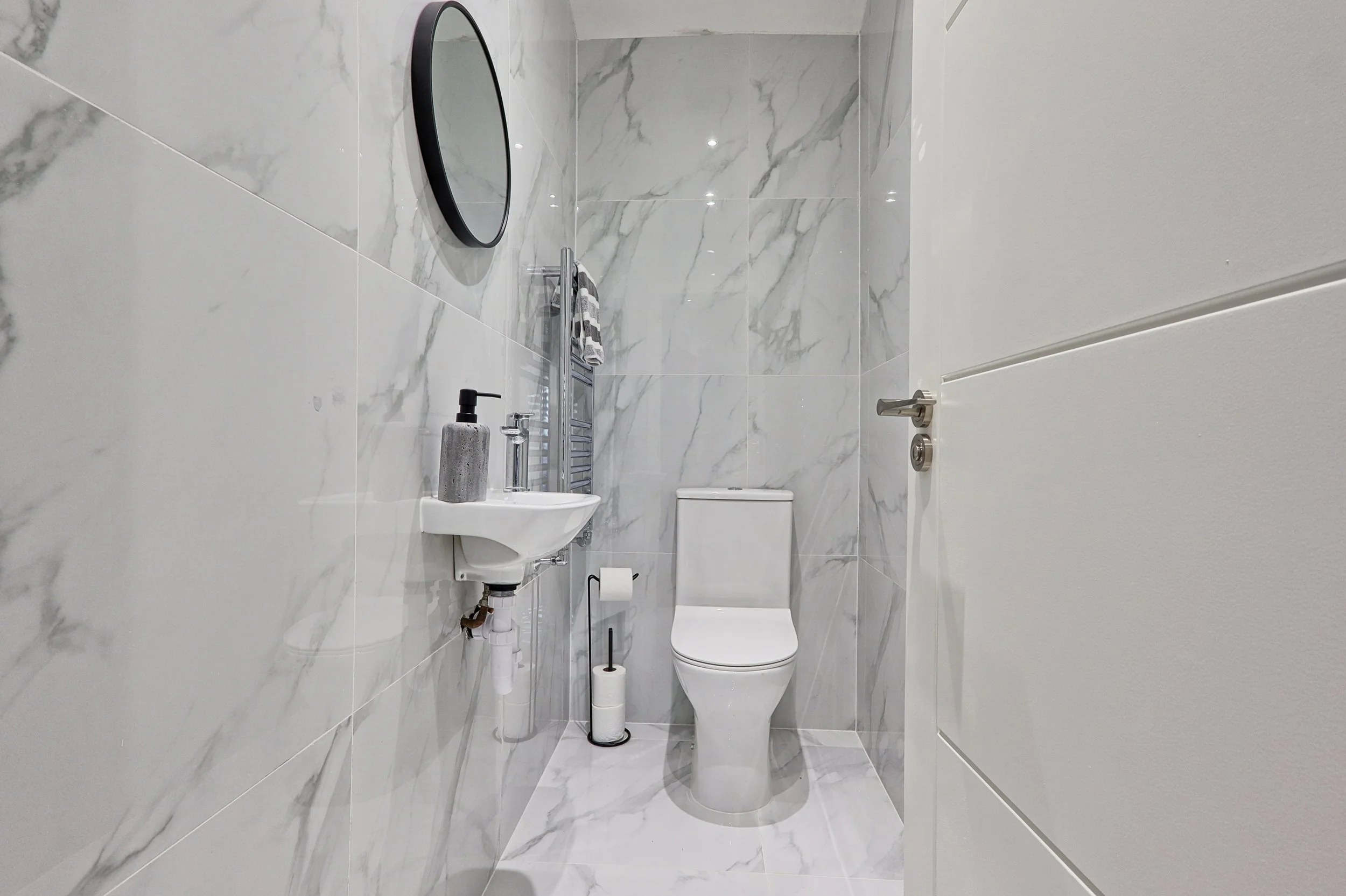 Small modern bathroom with marble walls and floor, a white toilet, a small wall-mounted sink with a grey soap dispenser, a wall-mounted mirror, and a toilet paper holder.