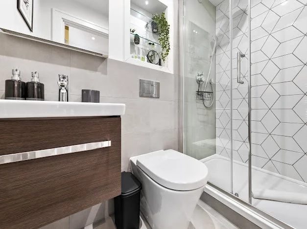 Modern bathroom with white tiles, a glass shower enclosure, a toilet, a wooden vanity, and a mirrored cabinet with plants and decorative items.