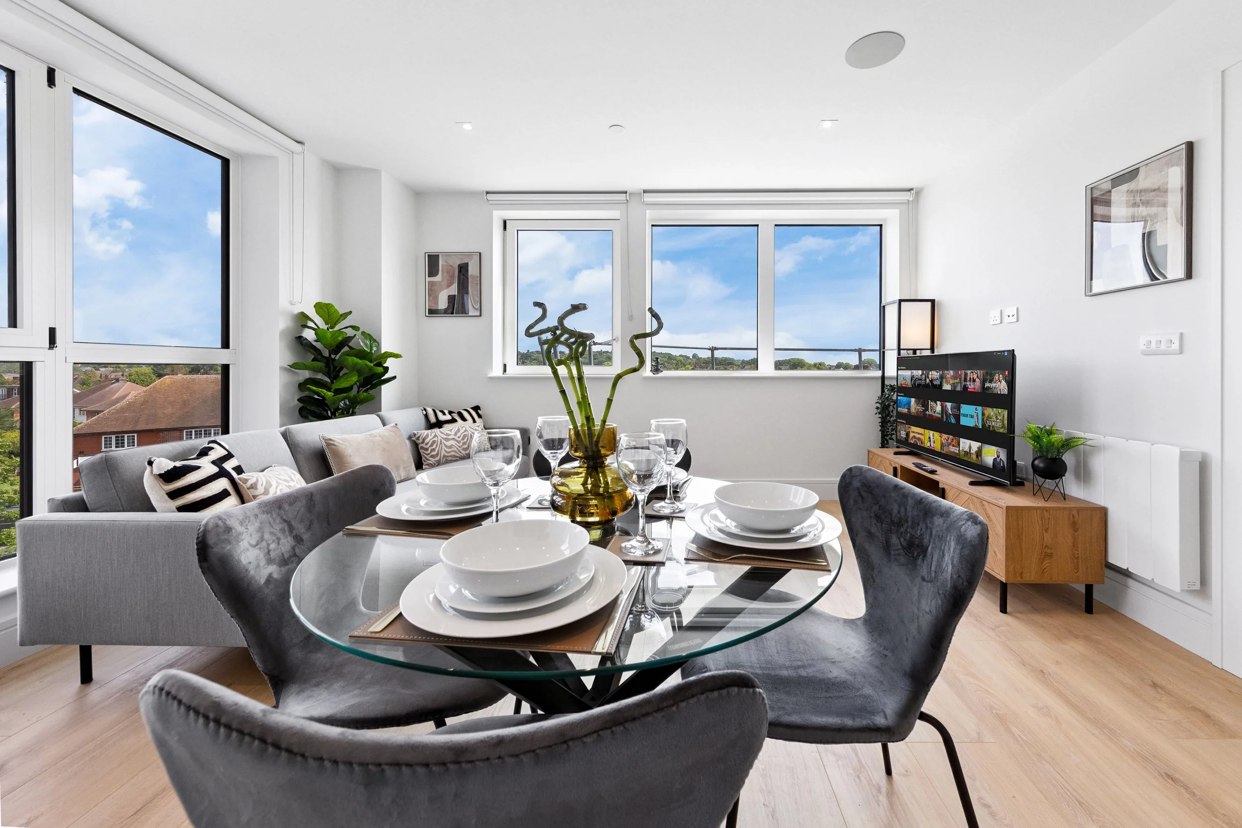 Modern living room with a round glass dining table set for four, grey velvet chairs, a grey sofa with decorative pillows, and a wooden TV stand with a flat-screen TV. Large windows offer a view of a suburban neighborhood with houses and trees.