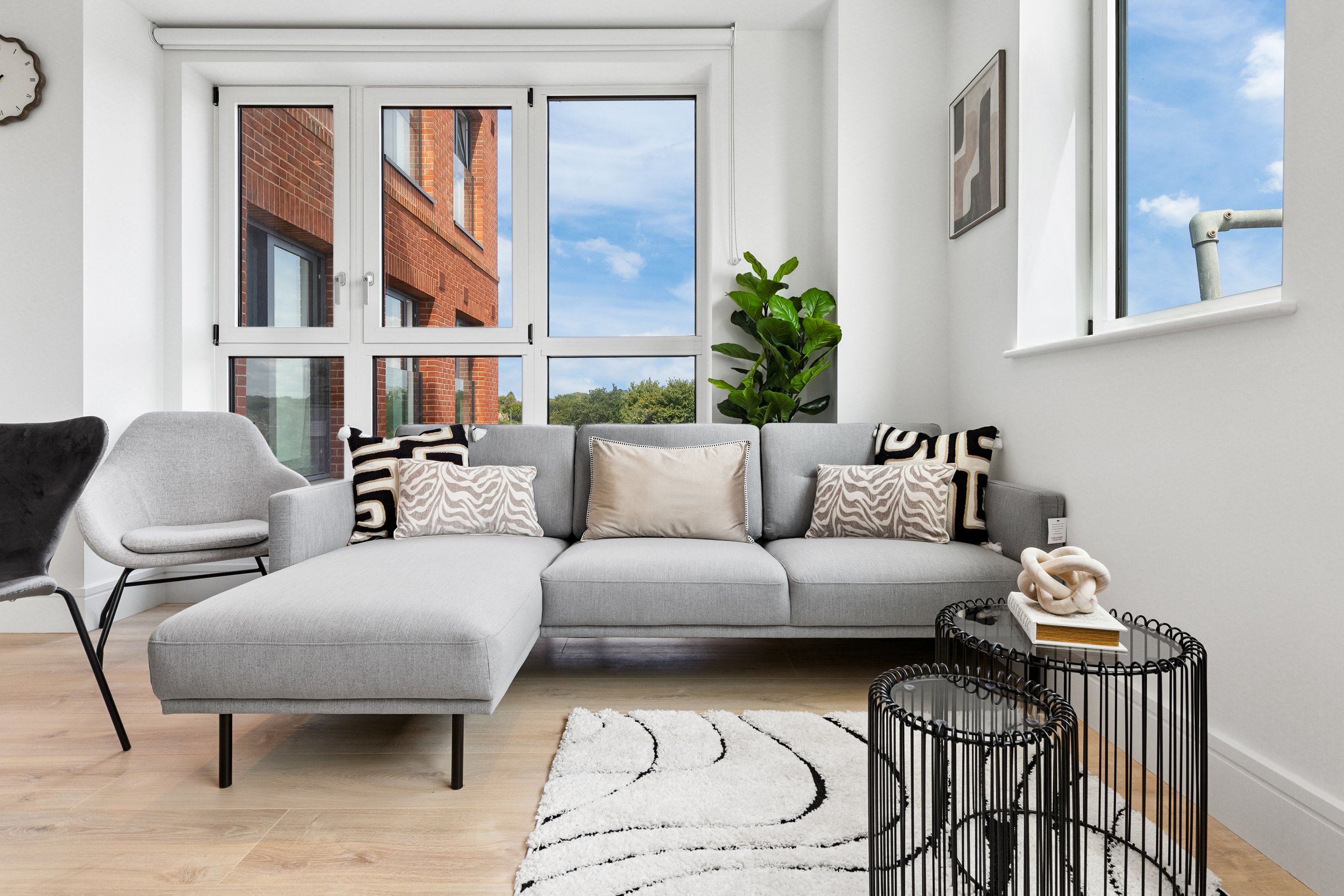 Modern living room with a large window, gray sectional sofa with decorative pillows, a white armchair, black wire side tables, a white shag rug, and a potted plant, with a view of a red brick building and blue sky outside.