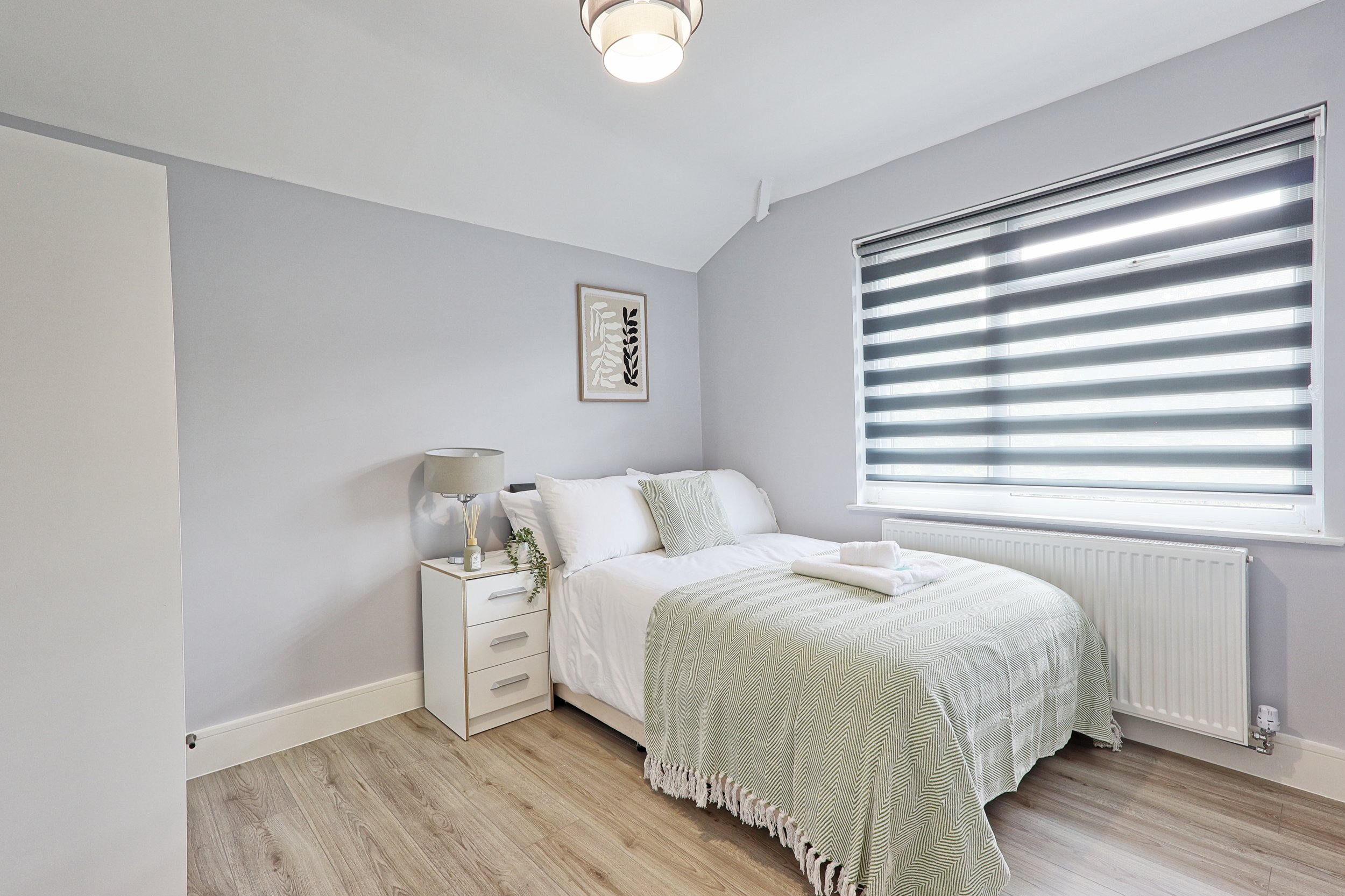 A clean, modern bedroom with a white bed, light green throw blanket, and a single pillow. There is a white nightstand with a gray lamp, some green decorative plants, and a framed artwork on the wall. The room has a large window with black and white striped blinds, a radiator below the window, and hardwood flooring.