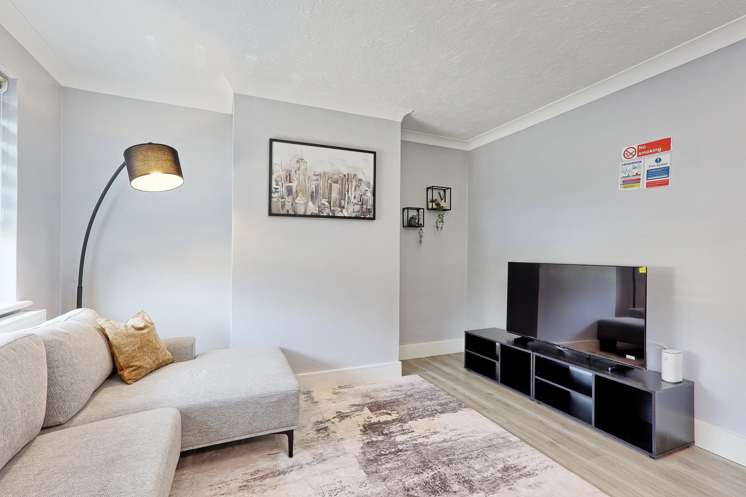 Living room with beige sectional sofa, throw pillow, black floor lamp, wall art, wall-mounted plants, flat-screen TV on black stand, and a no smoking sign on the wall.