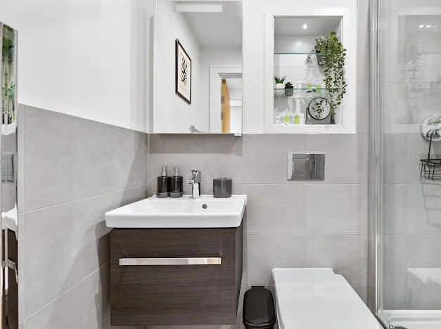 Modern bathroom with a white sink, dark vanity, gray tile walls, mirror, and a decorative shelf with plants and clock.