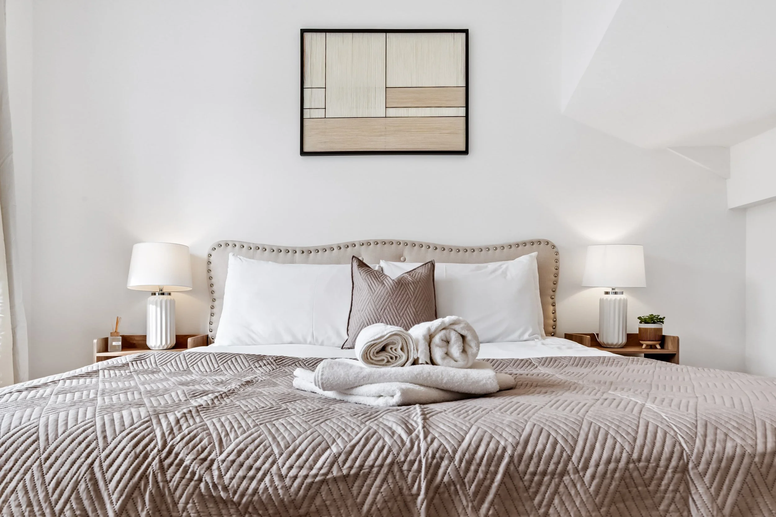 A neatly made bed with beige quilt, white pillows, a brown decorative pillow, and rolled white towels on top, in a bright bedroom with white walls, two bedside tables with lamps, and a framed geometric art piece above the headboard.