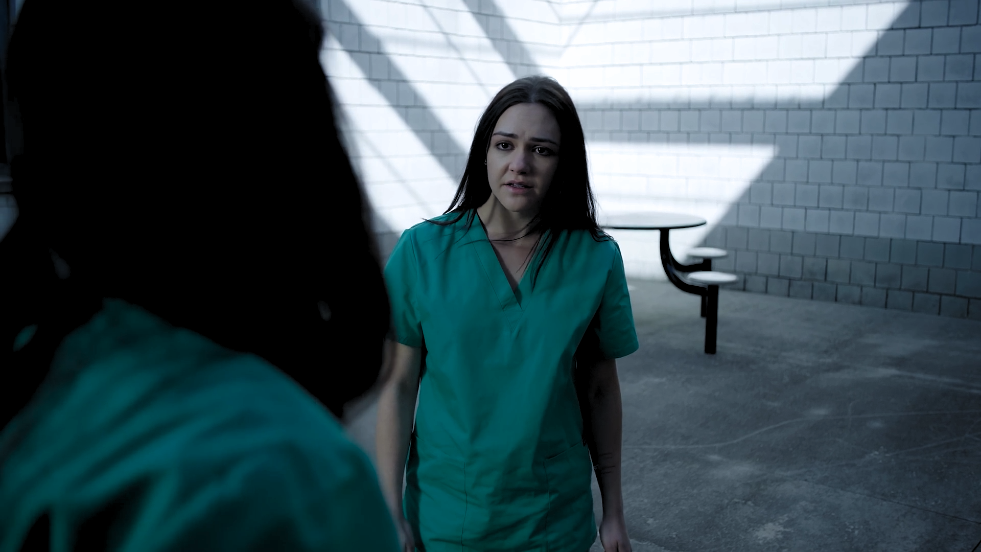 A woman in green scrubs with dark hair talking to a person with curly hair in the foreground, against a white brick wall and stair railing in the background.