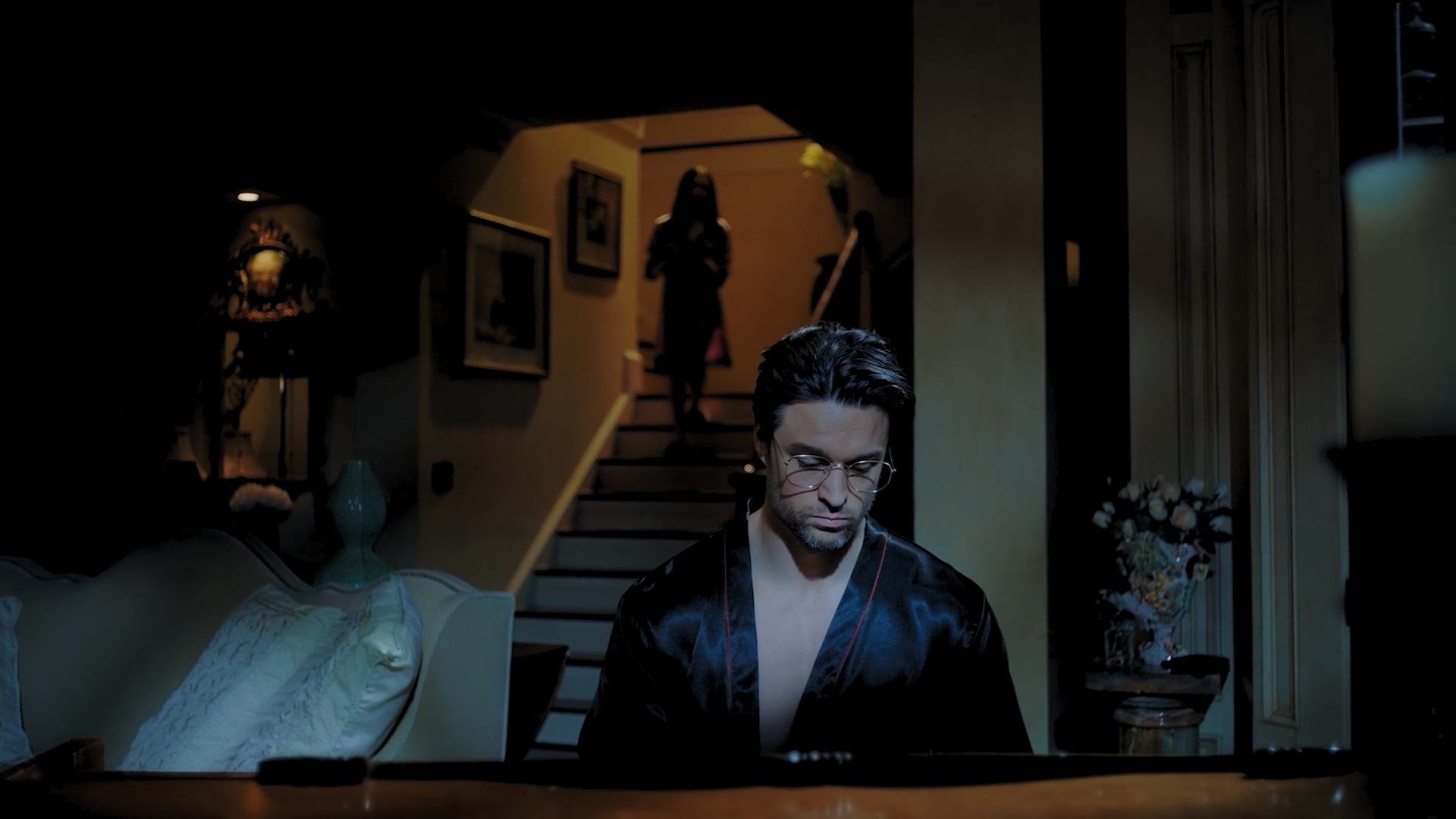 A man with glasses and dark hair sitting at a table, looking down, with a staircase and a woman standing at the top in the background inside a dimly lit home.