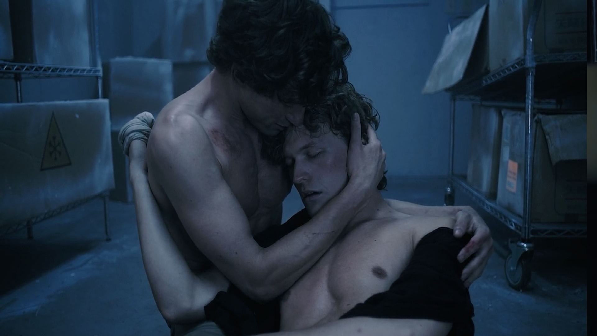 Two shirtless men in an emotional embrace in a dimly lit room, with metal shelving units and cardboard boxes in the background.