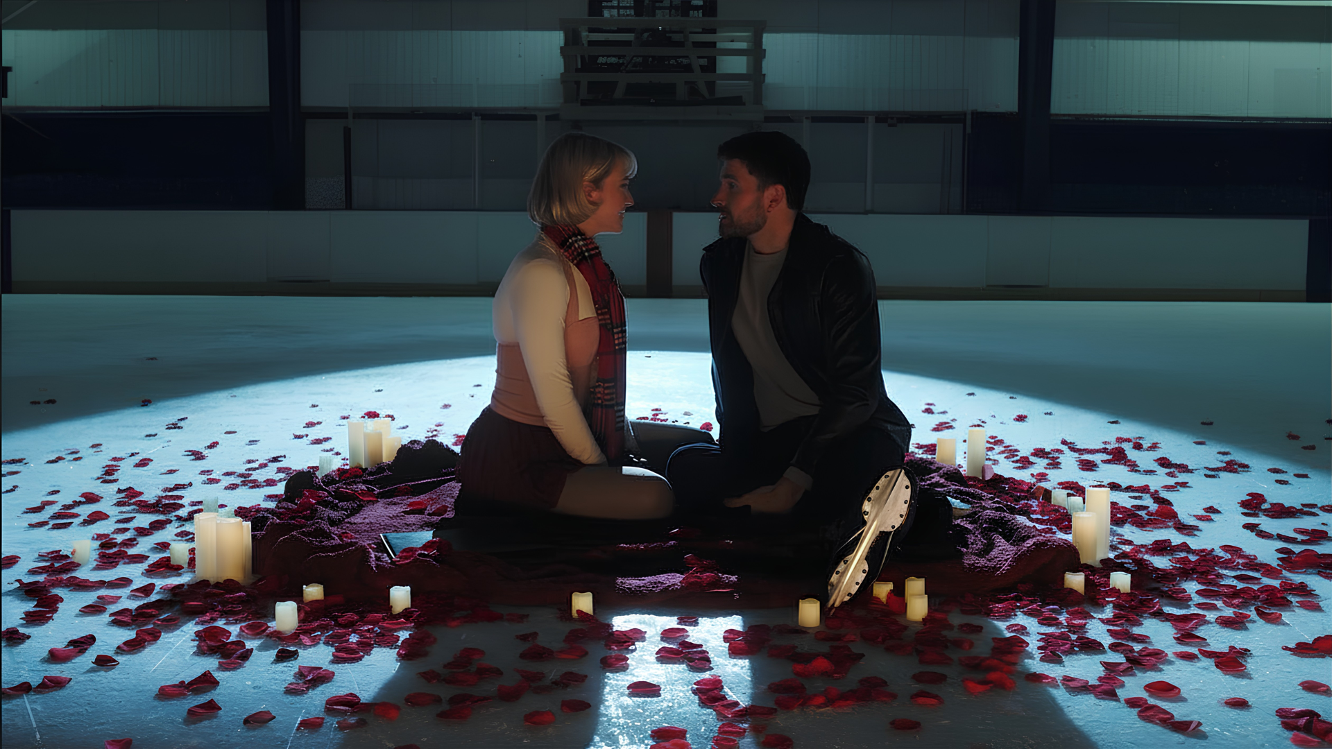 A couple sitting on the floor surrounded by candles and rose petals, facing each other in a romantic setting with dim lighting.