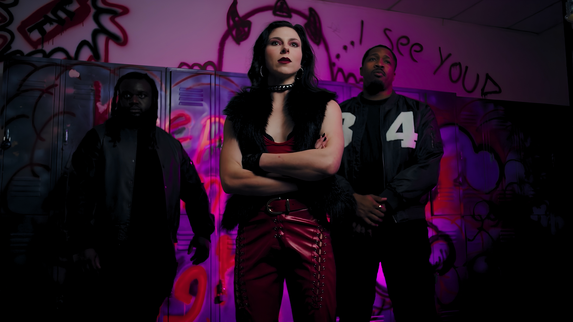Three people standing in front of garage lockers with graffiti and pink lighting, with a woman in the center wearing red pants with lace-up details and dark makeup, flanked by two men in black jackets.